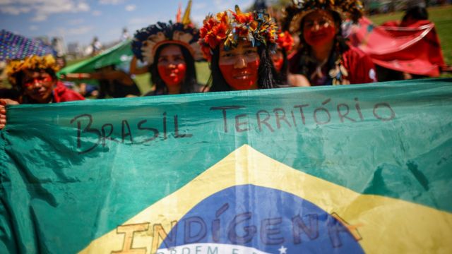 Jovens mulheres indígenas seguram bandeira do Brasil com os dizeres "Brasil Território Indígena"