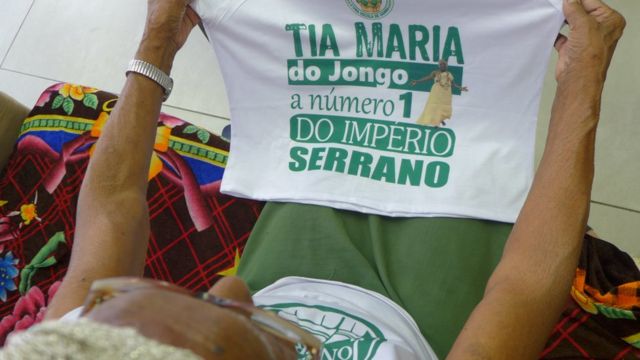 Tia Maria segurando a camiseta do Império Serrano que diz que ela é a número 1 da escola