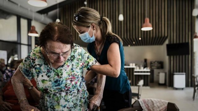A volunteer helps a woman standing over a chair at a table during lunch at the Landais Alzheimer site for Alzheimer's patients in Dax, France