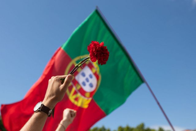 Mão segura cravo vermelho com bandeira de Portugal ao fundo