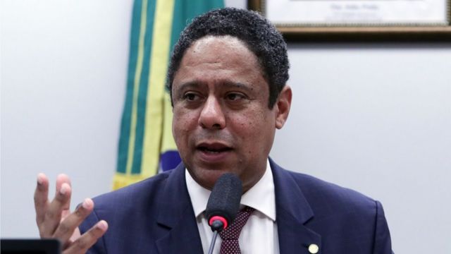 Retrato do deputado federal Orlando Silva (PC do B-SP) numa comissão da Câmara dos Deputados. Ele é um homem negro. Veste camisa social branca e gravata vinho com um terno azul marinho.