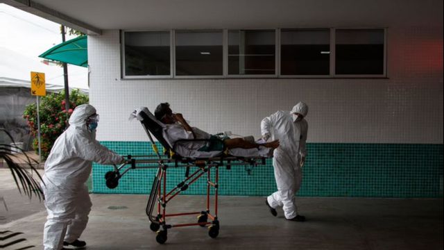 Duas pessoas paramentadas carregam paciente em maca em parte externa de hospital