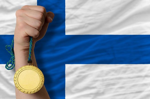 Braço branco segura uma medalha de ouro em frente a uma bandeiras da finlândia