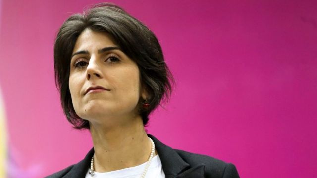 Manuela D'Ávila com olhar confiante, em pé em frente a painel rosa
