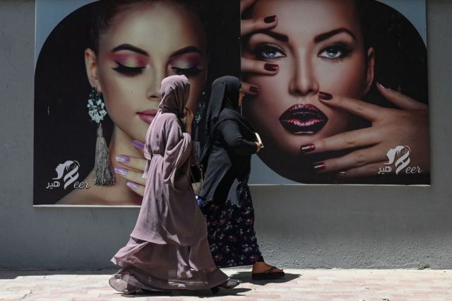 Mulheres vestidas de burca caminham na frente de retratos de outras mulheres maquiadas em Cabul, em 7 de agosto de 2021