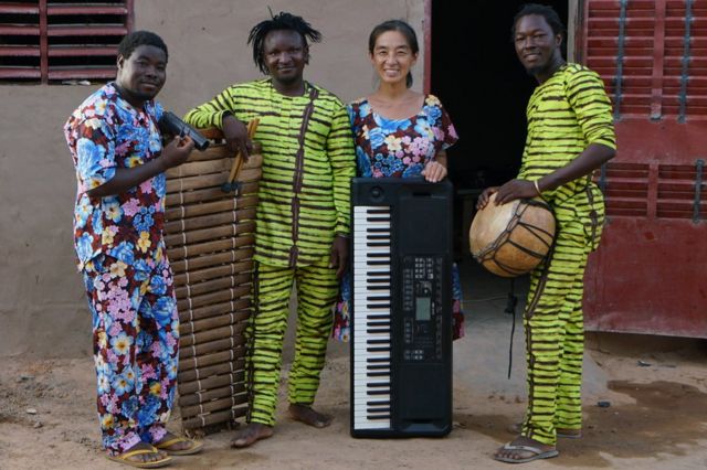Maboudou Sanou, Boureima Sanou, Keiko Fujiie et Ibrahim Dembélé posent pour une photo devant la maison que Fujiie partage avec Maboudou Sanou et sa famille à Ouagadougou. Les musiciens sont issus d'une famille de griots de la ville de Nouna, dans l'ouest du Burkina Faso, dans laquelle les traditions musicales ont été transmises de génération en génération. Le groupe répète du lundi au vendredi.