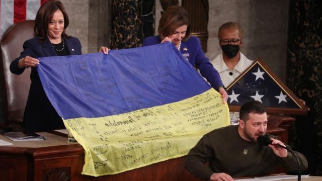 米議会で演説するゼレンスキー大統領（手前）と、ウクライナ兵が名前を書き込んだウクライナ国旗を手にしたカマラ・ハリス米副大統領（左）とナンシー・ペロシ下院議長（右奥）