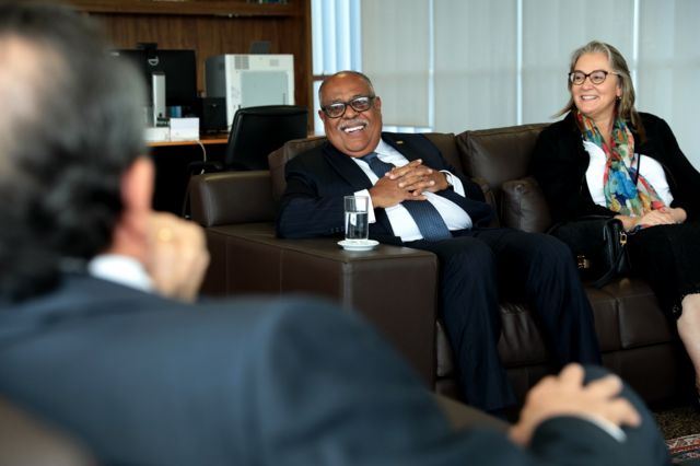 O ministro Benedito em seu gabinete no TSE. Ele está de terno azul escuro risca de giz e gravata azul, sentado em um sofá marrom com as mãos cruzadas sobre a barriga.