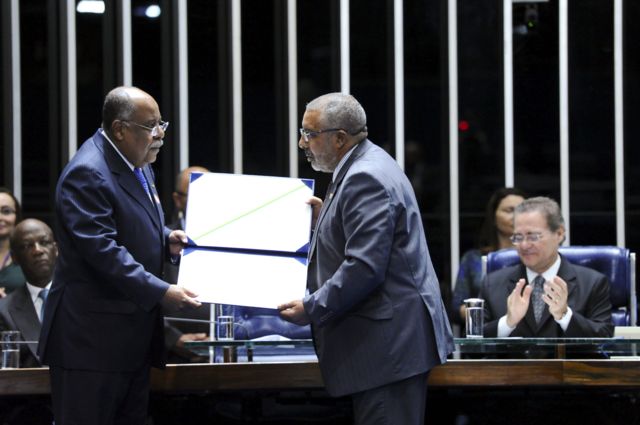 O ministro Benedito Gonçalves e o senador Paulo Paim no Plenário do Senado. Ambos são homens negros idosos de terno. Paim estende um diploma comemorativo a Benedito
