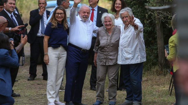 Lula, Mujica e suas esposas abraçadas e sorrindo para foto em área arborizada