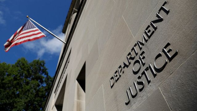 The Department of Justice symbol is seen on a wall beneath a flying American flag in this low-angle shot