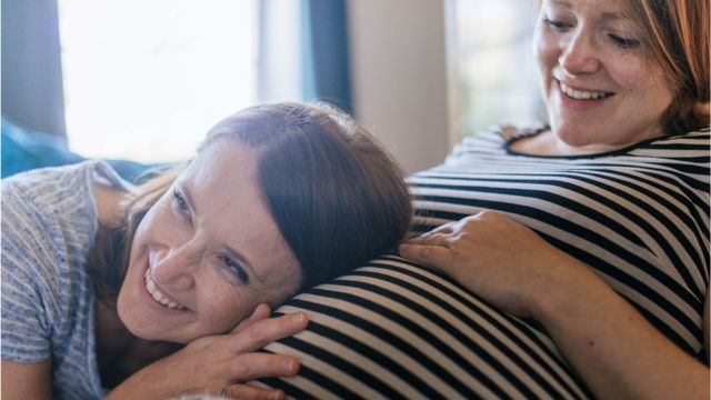 One woman puts her ear on the pregnant belly of another.