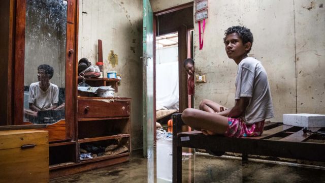Meninos em casa inundada em Fiji, com móveis e pertences danificados pela água