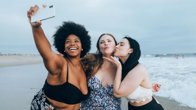 Imagem mostra três mulheres fazendo selfie em praia