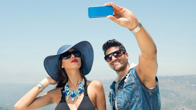 Man in sunglasses posing for a selfie in a sunny location with a lady in a a sunhat and bright red lipstick