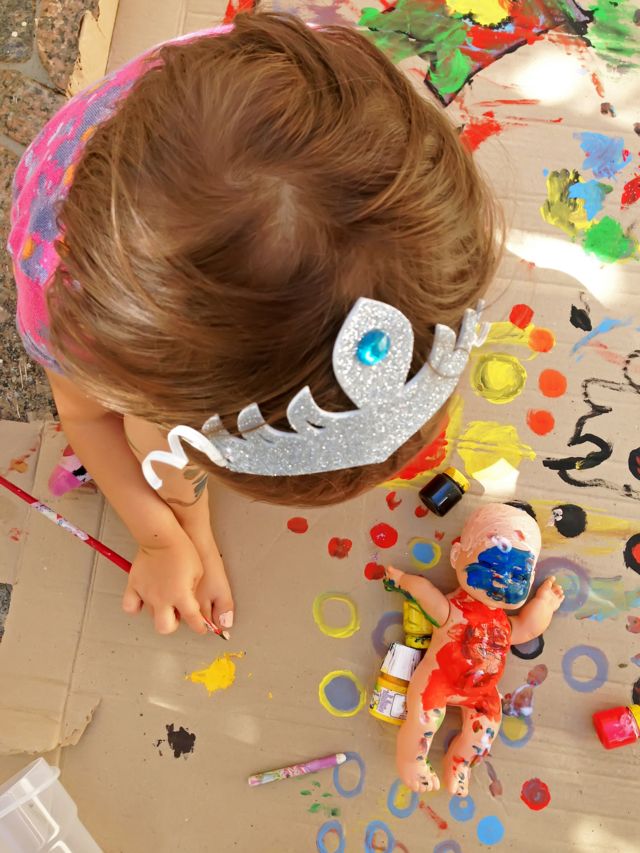 Menina com pincel na mão e boneca no chão com o corpo pintado com tintas coloridas
