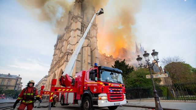 Bombeiro e caminhão aparecem em ação com a catedral pegando fogo ao fundo