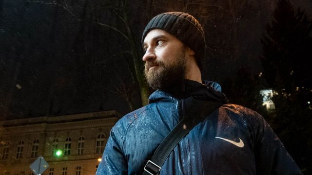 Fotografia colorida mostra homem jovem e branco de barba castanha, touca e roupas de frio sob um céu noturno de cidade