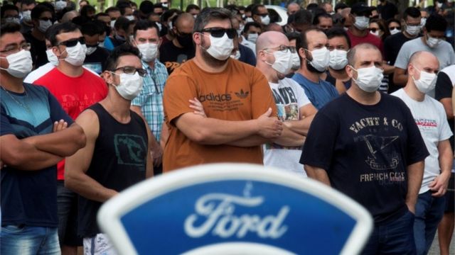 Dezenas de homens e mulheres de máscaras e braços cruzados durante protesto, com logo da Ford na frente destas pessoas