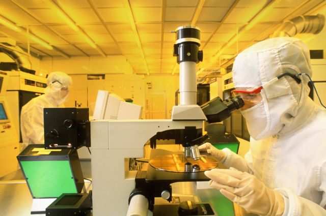 Técnico utilizando el microscopio para inspeccionar la oblea de silicio con chips de ordenador en la sala limpia de producción.