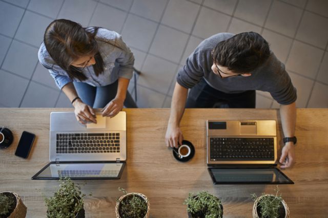 Homem e mulher jovens tomando café e conversando em frente aos respectivos laptops