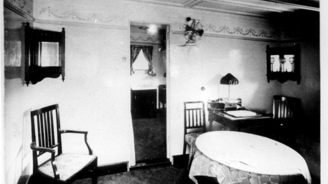 Foto em preto e branco de uma sala com cadeiras, mesa e um relógio