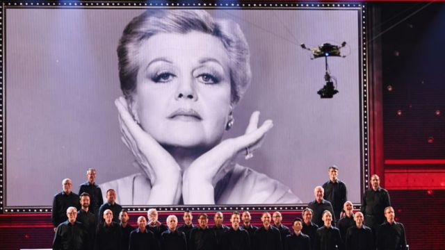 Un coro cantando con una imagen foto de Angela Lansbury proyectada atrás.