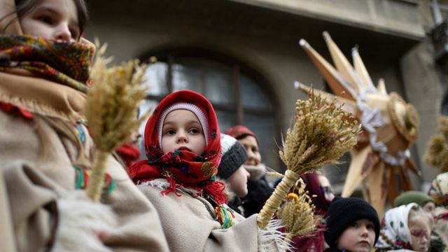 A Ukrainian girl in a red floral scarf holds a bundle of wheat during a Christmas ceremony.