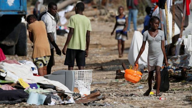 Una adolescente camina por una calle de Haití sosteniendo una cesta plástica y a su alrededor hay escombros y ropa tirada.