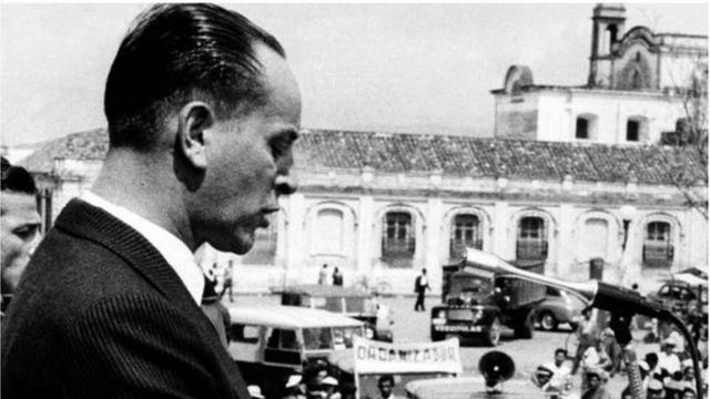 Jacobo Arbenz pronuncia un discurso frente a varias personas en un espacio público.