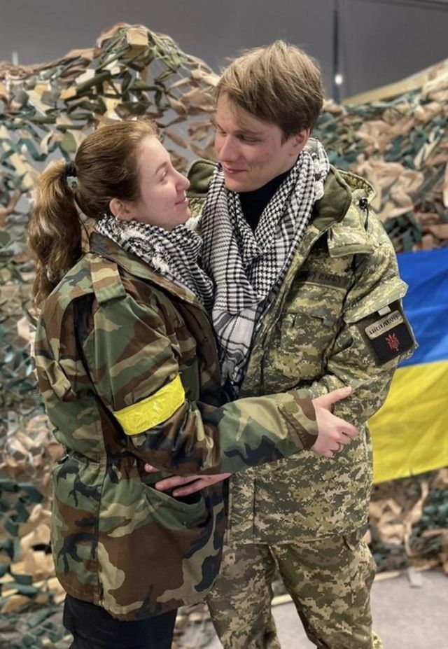 Yaryna Arieva and Svyatoslav Fursin hug each other in military uniforms with a Ukrainian flag behind them.