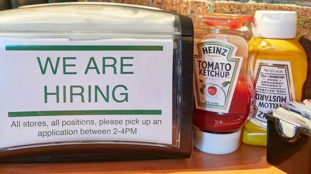 Cartaz de contratação de funcionários ao lado de bisnaga de ketchup e mostarda em balcão