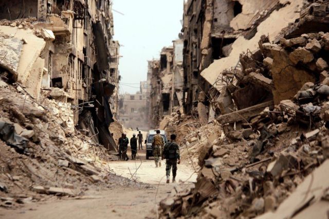 Para tentara berjalan melewati bangunan yang hancur di kamp Yarmouk Palestina di Damaskus, Suriah.