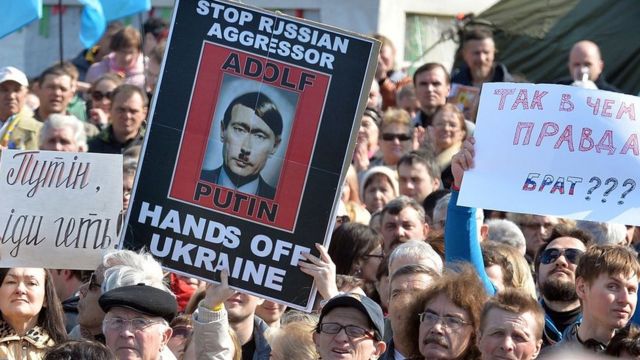 Manifestantes seguram cartaz que retrata o presidente russo, Vladimir Putin, como Adolf Hitler em que se lê: 'Detenham o agressor russo, tire as mãos da Ucrânia' — em março de 2014, em Kiev