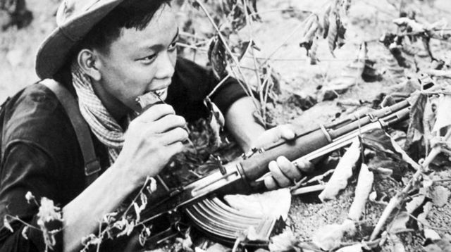 La photo en noir et blanc montre un soldat vietcong muni d'un fusil AK-47. Le soldat, légèrement de profil, semble être allongé sur le ventre sur le sol.