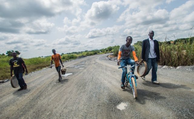 A group of men walk and cycle along a road with their equipment