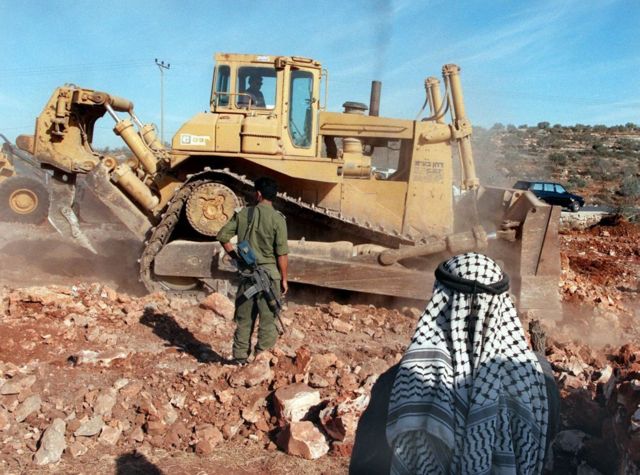 Un palestino contempla el trabajo de una excavadora custodiada por soldados israelíes