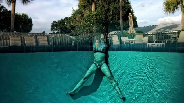 Mulher de biquíni numa piscina com parte do corpo submersa e parte fora da água criando um efeito ótico entre as duas partes