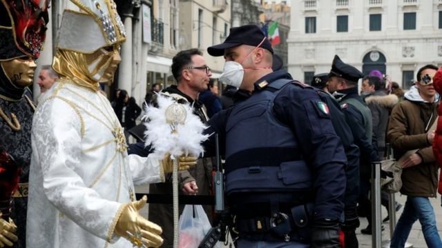 Police officer wearing a protective face mask stands next to carnival revellers at Venice Carnival. 23 Feb 2020