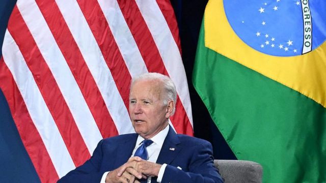 Presidente americano Joe Biden sentado em frente às bandeiras dos EUA e Brasil