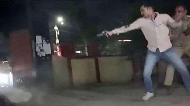 தாக்குதல் நடத்துபவரை பின்னால் இருந்து பிடிக்க முயற்சிக்கும் போலீஸ்காரர்.