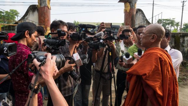 A Buddhist monk in orange robes addresses a gaggle of journalists on the monastery grounds in Maungdaw