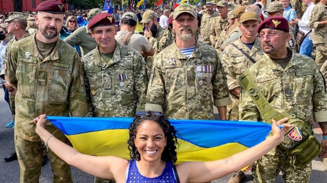 Fernanda Krupin segurando uma bandeira da Ucrânia em frente a homens fardados, durante la celebración de la independencia del país
