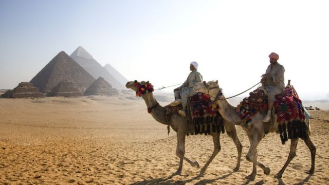 Dos hombres en camello con las pirámides de fondo.