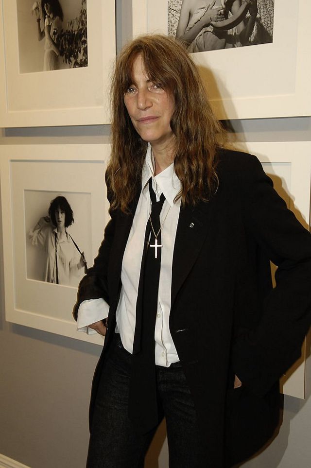 Пэтти Смит в таком же черном пиджаке и таком же черном галстуке на выставке Роберта Мапплторпа перед сделанными им в 1970-е годы ее портретами