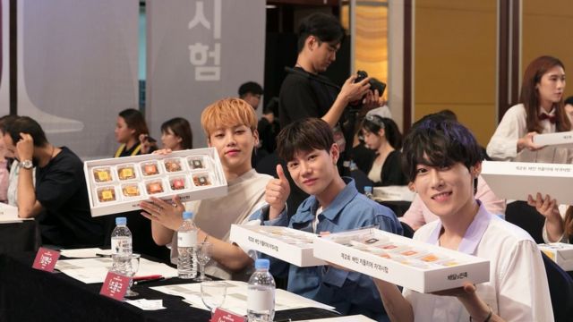 Three South Korean young men give thumbs up and show boxes with ten pieces of fried chicken for blind tasting