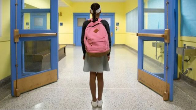A girl with a school bag in school uniform stands in and empty corridor with her back to camera