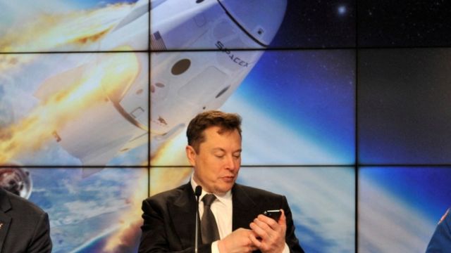 Musk olha para celular, com imagem de foguete na tela atrás