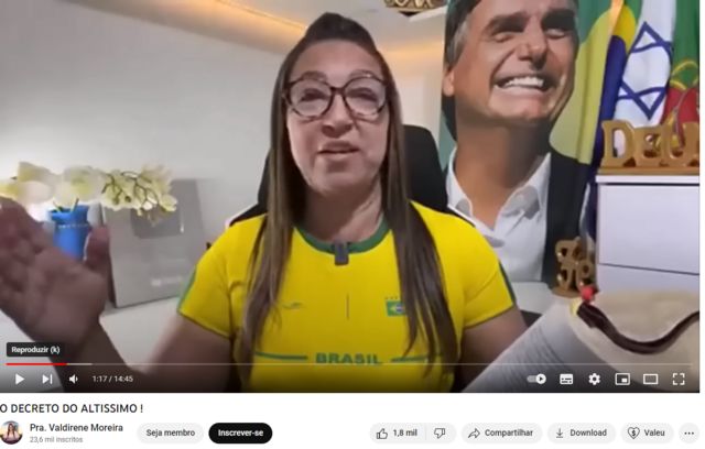 Valdirene Moreira sentada em cenário com bandeiras do Brasil, Portugal e Israel ao fundo, além de pôster com foto de Bolsonaro e placa comemorativa que ganhou do YouTube