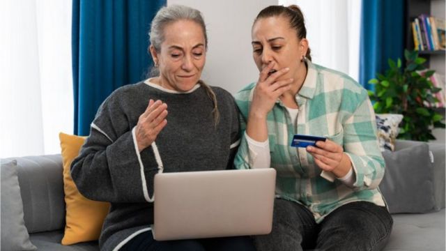 Duas mulheres sentadas no sofá — uma delas com o laptop no colo, e a outra com cartão de crédito na mão e cara de assutada